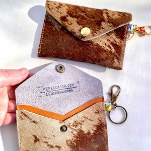 May include: Two cowhide leather cardholders. One is closed with a gold snap, the other is open, revealing an orange interior and the text "REBECCA HALCOM LEATHERWORKS". Both have keychains with gold clasps.