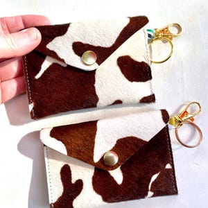 May include: Two cowhide cardholders with a brown and white pattern. Each has a gold-colored clasp and a snap closure. One is held by a hand. The cardholders are small and rectangular.