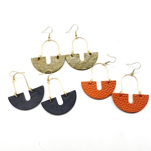 May include: A collection of handmade earrings in various colors. The earrings feature a gold-colored metal arch and a semi-circular leather base. Colors include olive green, navy blue, and orange. The earrings are designed for pierced ears.