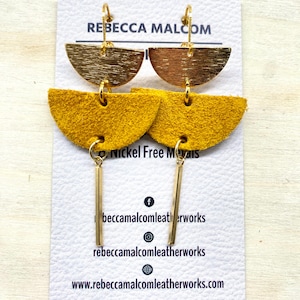 May include: Gold-toned metal and mustard yellow leather earrings. The earrings feature a half-circle design with a gold bar pendant. The card behind the earrings reads "REBECCA MALCOM" and "Nickel Free Metals".