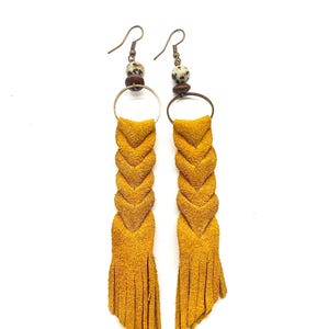 Puede incluir: Un par de pendientes de gamuza amarilla mostaza con un diseño trenzado y flecos. Los pendientes tienen una pequeña cuenta de madera marrón y una cuenta de piedra moteada.