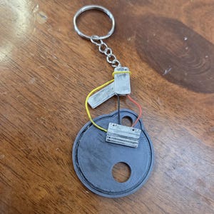 May include: A custom-made keychain with a circular, dark gray base and a silver key ring. The keychain features a small electronic component with yellow and red wires, and a silver metal piece.