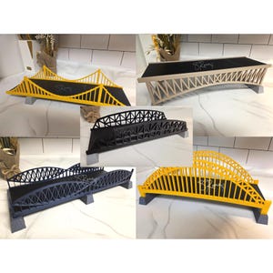 May include: A collection of five bridge models featuring various structural designs and colors including yellow, beige, black, and dark blue. Each model has a flat black deck with the text S Rona printed on it and rests on small gray supports.