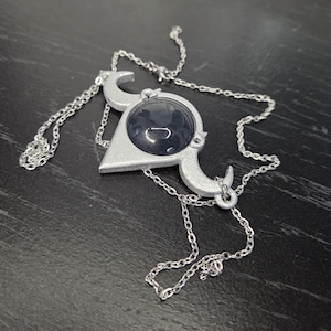 May include: Silver-toned pendant necklace with a geometric design. The pendant features a dark, round center stone set within a silver frame with crescent moon accents. The necklace chain is delicate and silver, resting on a dark surface.