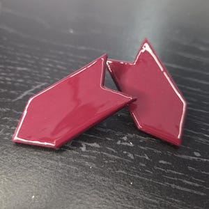 May include: Two burgundy-coloured, arrow-shaped objects with a glossy finish and white trim. The objects are positioned on a dark surface, with one slightly overlapping the other. The design is geometric.