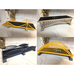 May include: Four miniature bridge models in yellow, beige, and dark blue, each with a black surface and gray supports. The bridges have intricate designs and are displayed on a white surface. Each bridge has the signature "83Rony" on the black surface.