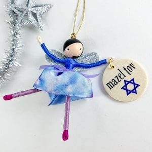 May include: A blue fairy ornament with silver glitter wings and a blue and white tie-dye skirt. The ornament has a round tag that says "mazel tov" with a blue Star of David.