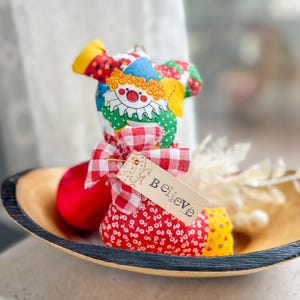 May include: A colorful handmade clown doll with a red, white, and blue patterned hat, a red nose, and a red and white checkered bow. The doll is holding a tag that says "Believe". It is placed in a wooden bowl.