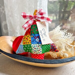 May include: A handmade fabric Christmas tree with a patchwork design in various colors, topped with a red and white gingham bow. A small tag with the words "Cozy Home" hangs from the tree. The tree sits in a wooden bowl with a red ornament.