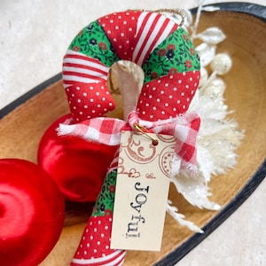 May include: A festive Christmas candy cane decoration made from fabric with red and white patterns. It features a tag with the word "Joyful!" and a red and white gingham ribbon. A red bauble is placed next to the candy cane.