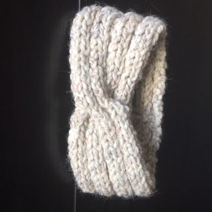 May include: A white knitted headband with a twisted design. The headband is made of a soft, chunky yarn.