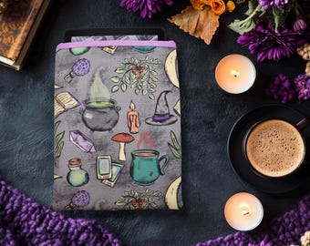May include: A gray tablet sleeve with a witchy theme, featuring illustrations of a cauldron, crystals, a witch's hat, and a mug. The sleeve is surrounded by purple flowers, candles, and a cup of coffee.