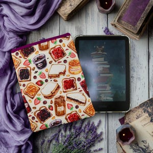 May include: A white and cream Kindle cover with a colorful pattern of peanut butter and jelly sandwiches, strawberries, and jars of peanut butter and jelly. The cover is on a white wooden surface with a Kindle e-reader next to it.