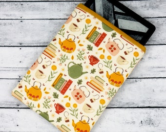 Puede incluir: Una funda para lector electrónico de color crema con un ribete amarillo mostaza. La funda presenta un patrón repetido de teteras, tazas de té, libros y flores en tonos de verde, amarillo, naranja y rosa.