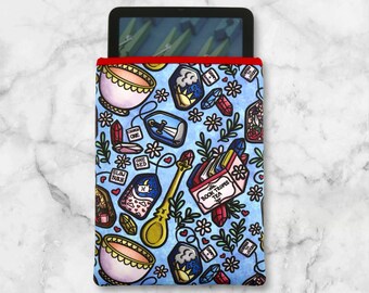 May include: A blue and white patterned e-reader sleeve with a red trim. The design includes teacups, tea tags, crystals, and a golden spoon. The sleeve holds a black e-reader.