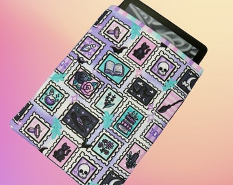 Könnte beinhalten: Eine E-Reader-Hülle mit einem lila, rosa und blauen Muster, das verschiedene hexenhafte und magische Symbole zeigt, darunter eine Katze, einen Schädel, ein Buch, eine Zaubertrankflasche, einen Besen und einen Halbmond.