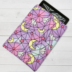 May include: Purple tablet sleeve with a geometric pattern of moons, stars, and bunnies. The pattern is in shades of pink, yellow, and white on a purple background.