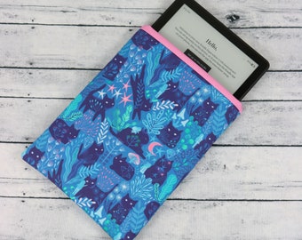 May include: A blue and pink e-reader sleeve with a black cat and floral pattern. The sleeve is made of fabric and has a pink zipper. The e-reader is inside the sleeve and the screen shows the welcome message "Hello".