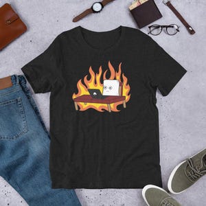 Puede incluir: Camiseta gris oscuro con un personaje de dibujos animados de malvavisco sentado en un escritorio con un portátil, rodeado de llamas. El diseño incluye un escritorio, una silla y llamas en tonos naranja y amarillo. La camiseta es de manga corta.
