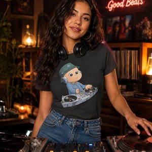 May include: A dark gray t-shirt featuring a cartoon graphic of a baby wearing a blue jacket, a gold chain, and a green bucket hat while operating a silver DJ turntable console. The baby has large eyes and is depicted with headphones around its neck.
