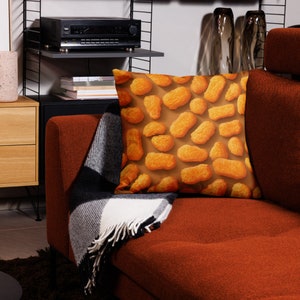 May include: A square throw pillow featuring a repeating pattern of golden-brown chicken nuggets on a brown background. The pillow is placed on a rust-colored sofa, with a black and white blanket draped over the arm.