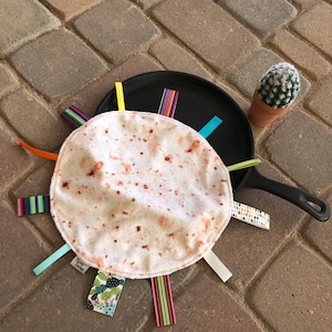 May include: A white tortilla-shaped baby toy with colorful fabric strips attached. The toy is lying on a black pan with a small cactus plant in a terracotta pot in the background.