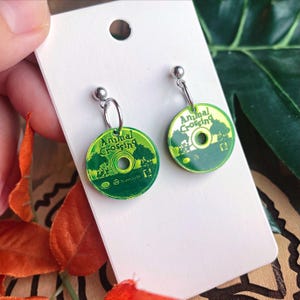 May include: A pair of green and yellow Animal Crossing earrings. Each earring is a small, circular disc with the game's logo and a tree design. The earrings have silver-colored hooks and posts.
