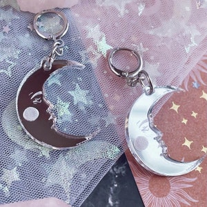 May include: Two crescent moon charms with a silver-tone metal hoop clasp. One charm features a dark brown reflective surface, while the other has a bright silver reflective surface. Both charms display a detailed face with a circular eye.