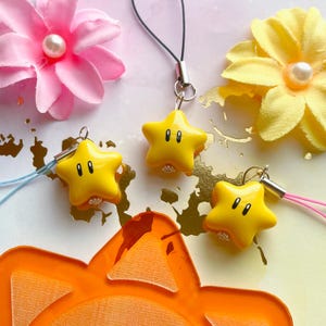 Puede incluir: Primer plano de amuletos amarillos en forma de estrella con ojos negros y cierres plateados. Cada amuleto está unido a un cordón de color. Los amuletos están colocados sobre un fondo de flores rosas y amarillas y un recorte naranja.