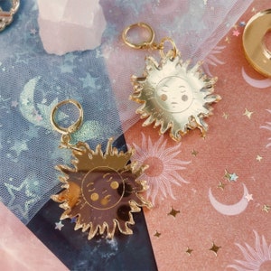 May include: Two sun-shaped charms with gold-toned mirror finishes, each featuring a stylized face with closed eyes and circular cheeks. The charms are attached to small gold-toned metal hoop clasps, designed for use as earrings or decorative pendants.