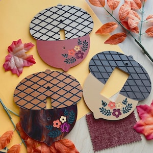 May include: Three acorn-shaped light switch covers featuring a textured grid pattern on the top half and floral designs on the bottom half. The covers are finished in maroon, brown marbled, and cream colors with small cutouts for switches.
