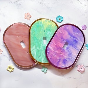 May include: Three oval light switch plates with a marbled finish in shades of dusty rose, mint green, and violet. Each plate features a central rectangular cutout for a toggle switch and two small circular holes for mounting screws.