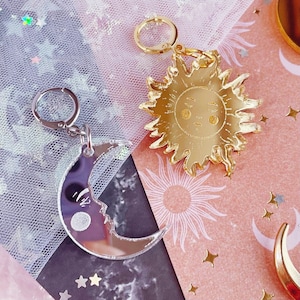 May include: Two charms with circular clasps: one silver-toned crescent moon with a face and one gold-toned sun with a face. The moon charm has a reflective surface, while the sun charm features a polished gold finish with detailed facial features.