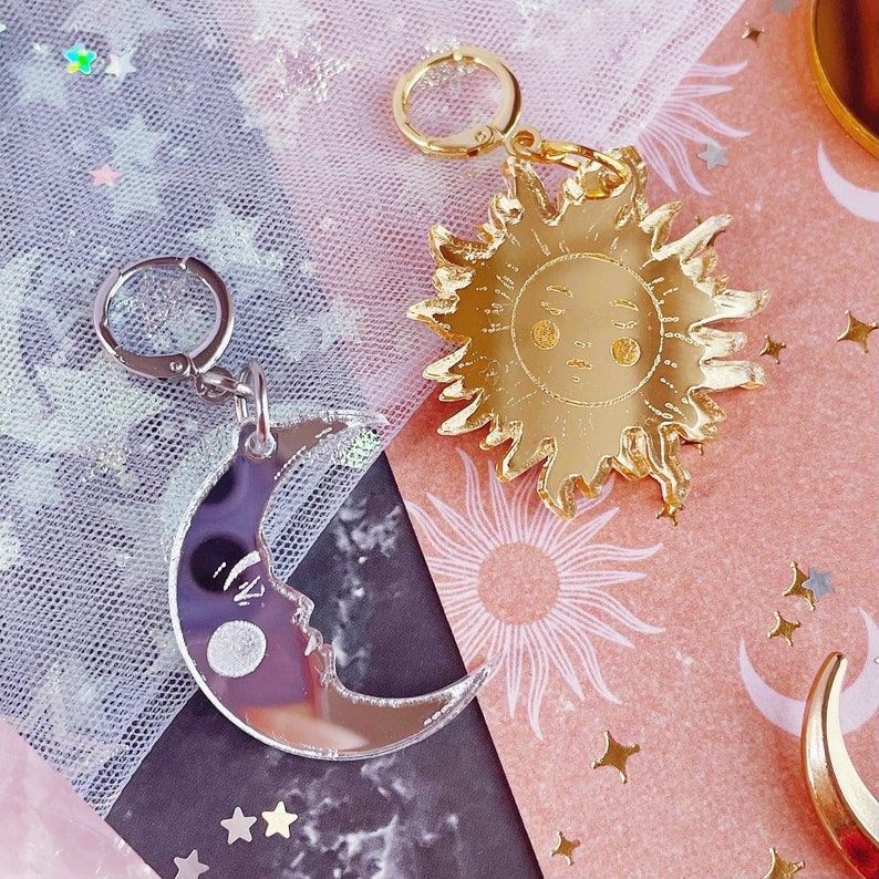 May include: Two charms with circular clasps: one silver-toned crescent moon with a face and one gold-toned sun with a face. The moon charm has a reflective surface, while the sun charm features a polished gold finish with detailed facial features.