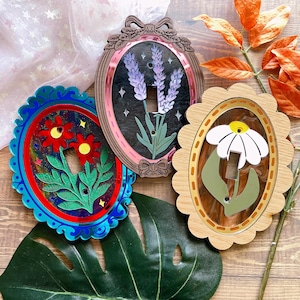 以下が含まれることがあります： 花柄のデザインが施された装飾的なライトスイッチカバー。赤い花、ラベンダー、デイジーがそれぞれ描かれています。各カバーはユニークなフレーム形状と配色を持ち、どんな部屋にも芸術的なタッチを加えます。カバーはアクリル製です。