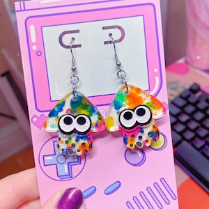 May include: A pair of colorful, iridescent earrings shaped like a squid or octopus. The earrings are made of clear acrylic with a rainbow paint splatter design. The earrings have silver hooks.