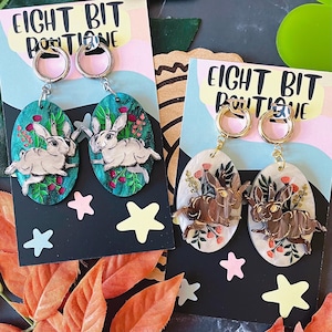 May include: Two pairs of oval-shaped dangle earrings attached to Eight Bit Boutique display cards. One pair features white rabbits on a teal background with floral accents, while the other pair shows brown rabbits on a cream background with flowers.