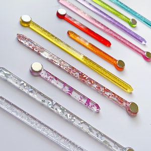 Puede incluir: Un conjunto de palitos acrílicos transparentes, brillantes y coloridos con extremos magnéticos. Los palitos tienen diferentes longitudes y presentan diferentes colores y patrones de purpurina.