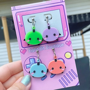 May include: Two pairs of dangle earrings featuring circular, flat, cartoon-style characters in lime green, lavender, light blue, and peach. Each charm has black eyes, pink cheeks, and small limbs, attached to silver-tone posts on a pink display card.