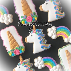 May include: A set of six unicorn and rainbow sugar cookies decorated with colorful frosting and gold accents. The cookies are arranged on a black background. The text "Shari's Cookie Shop" is visible in the image.