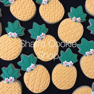 May include: A collection of yellow pineapple-shaped cookies decorated with green leaves and white flowers. The cookies are arranged on a black background. The text "Shari's Cookie Shop" is visible in the center of the image.