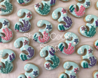 May include: Cookies shaped like the number 3, decorated with light blue icing and white polka dots. Each cookie features coral and starfish designs in pink, teal, and purple. The text "Shari's Cookie Shop" is visible.