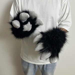 May include: A pair of black faux fur gloves designed as animal paws. Each glove features long, shaggy black synthetic fur covering the back of the hand and four padded, light gray fabric claws extending from the fingertips and palm area.