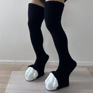 May include: A pair of black fuzzy over-the-knee socks featuring white plush animal paw designs at the toes. The socks have a soft, textured material that extends up the leg, ending above the knee, with padded white toe sections resembling claws.