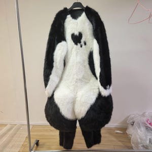 May include: A black and white panda costume made of faux fur, hanging on a black hanger. The costume features a heart-shaped design on the back. The costume includes a hood, body, and legs. The costume is designed to be worn.