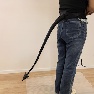 May include: A black, flexible, long, curved tail accessory attached to a black waist belt. The tail features a smooth, solid black finish and terminates in a pointed, triangular, arrow-shaped tip. The belt is secured around the waist.