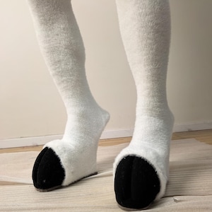 May include: A pair of knee-high white fuzzy socks featuring black hoof-shaped toe sections. The socks have a soft, textured appearance and are designed to mimic the look of animal legs with distinct black hooves at the base of each foot.