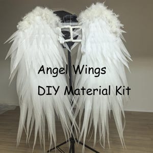 May include: White angel wings made of feathers and fabric, with the text "Angel Wings DIY Material Kit". The wings are displayed on a black stand, with a light-colored background.
