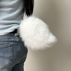 Peut inclure: Un accessoire de queue en fausse fourrure blanche et duveteuse fixé à une ceinture noire. La queue est épaisse, arrondie et composée de longues fibres synthétiques douces. Elle est placée au bas du dos, dépassant d'un jean en denim bleu.