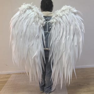 May include: Large white angel wings with a feathery texture. The wings are attached to a harness and worn on the back. The wings are made of white feathers and are a costume accessory.